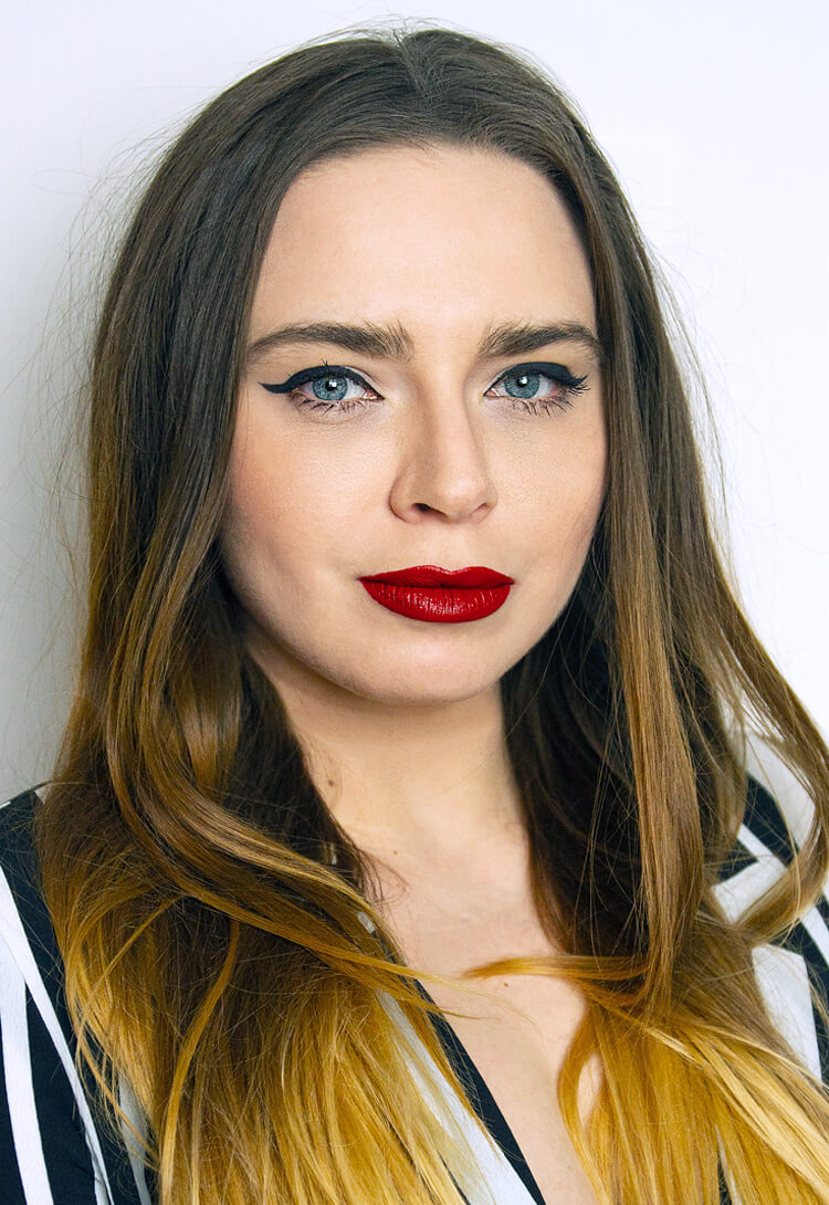 Self-portrait: dark hair, red lipstick, a striped top, against pale grey
