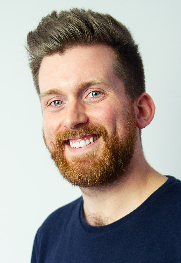A man with a short beard in a navy t-shirt, smiling, against pale grey