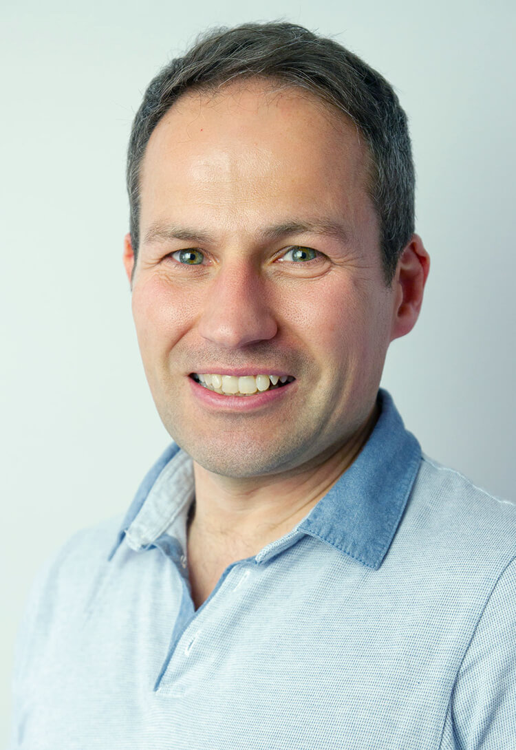 A man with greying hair in a pale blue polo shirt, smiling