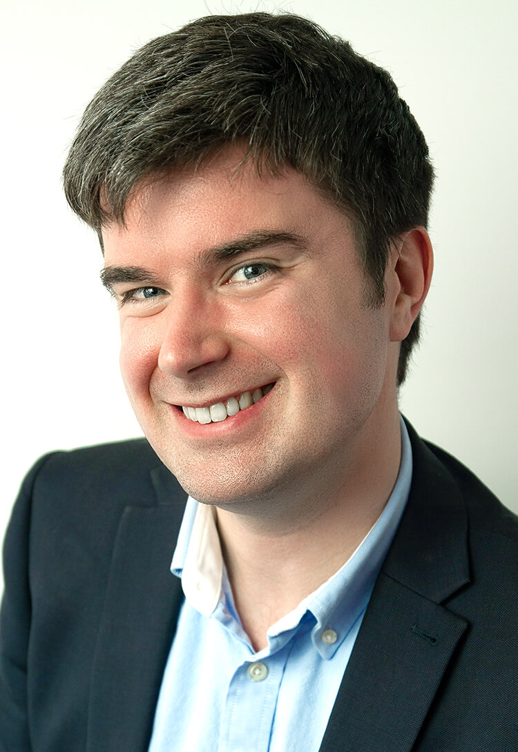 A man in a dark jacket and pale blue shirt, smiling