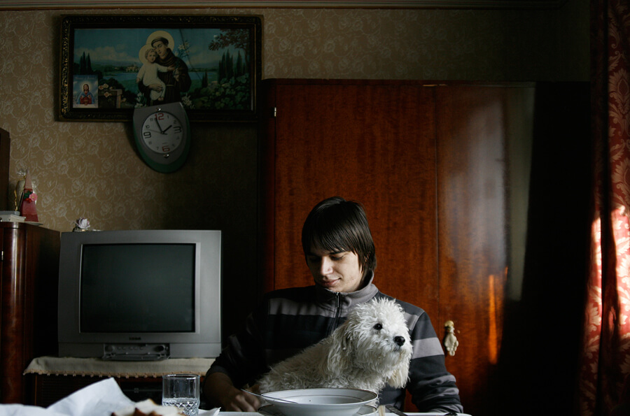 A young person at a table in a patterned room, holding a small white dog, an old television beside them
