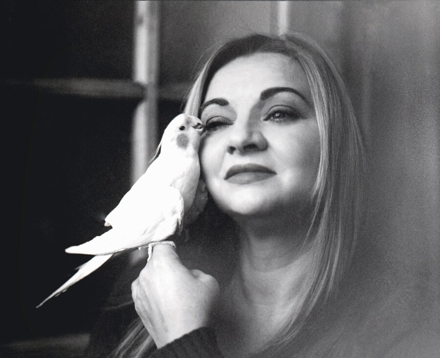A woman in black and white with a white cockatoo perched on her hand at her cheek
