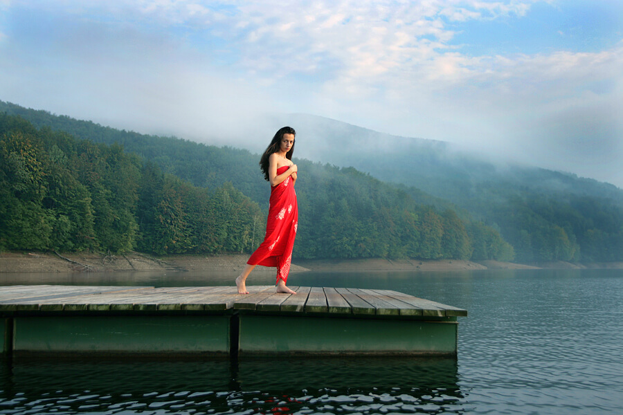 A woman in a red dress walking along a wooden jetty on a misty lake