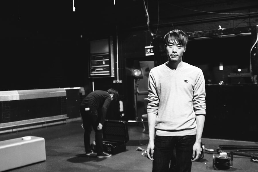 A young man standing in a dark studio while a crew member works behind him, in black and white