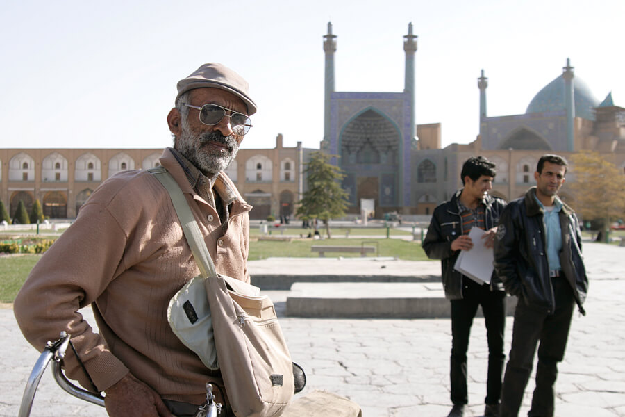A man in a flat cap and sunglasses in a square in Isfahan, the mosque domes behind him
