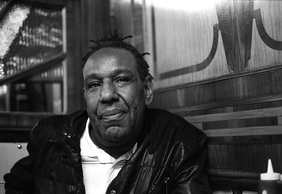 A man in a leather jacket outside a boarded-up shopfront, in black and white