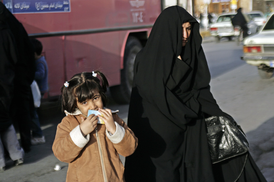 A small girl in a tan coat eating, a woman in a black chador beside her at the roadside