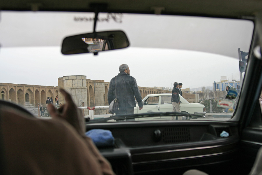 Through a car windscreen, a man standing on the bonnet of a parked car, an arcaded bridge behind