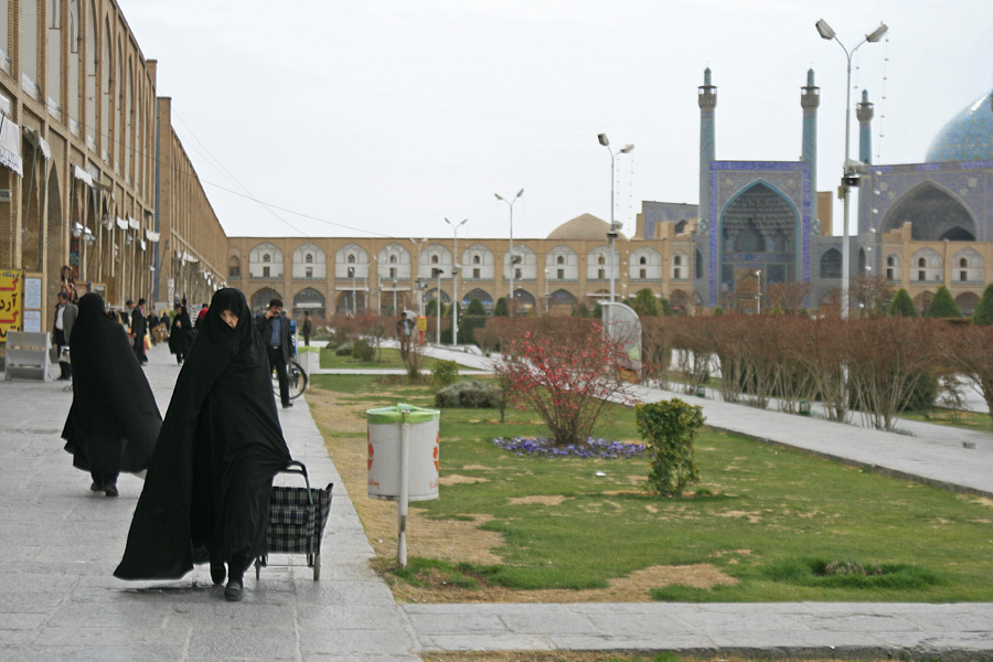 A public square with lawns, women in black chadors walking, a domed tiled mosque beyond
