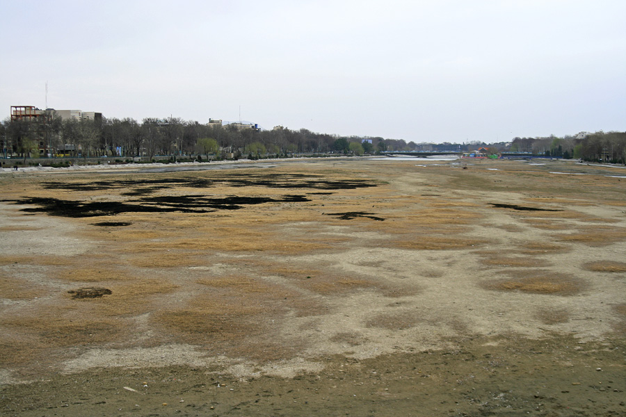 The same riverbed, dry and cracked, a line of trees along the far bank