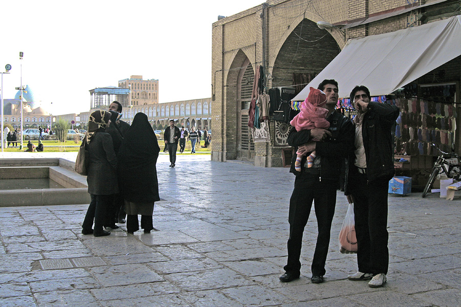 A couple holding a child in a square, a group of women in chadors nearby, market stalls behind