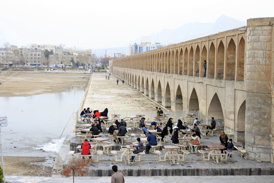People sitting at tables on the riverbank beneath the arches of a long stone bridge