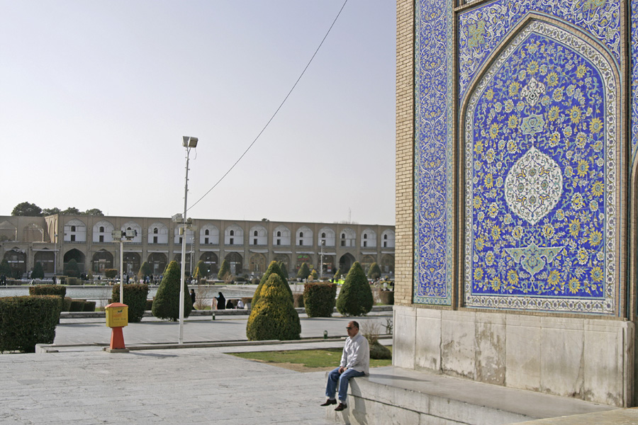 A tiled mosque portal at the edge of a large paved square, a man sitting on the low wall