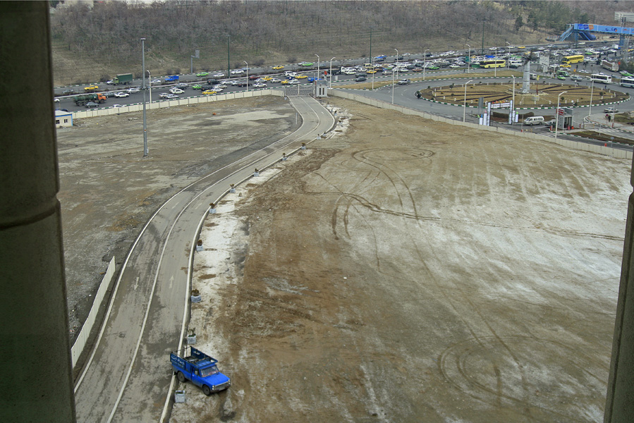A wide dry riverbed seen from above, a small blue truck on the sand, cars parked along the road
