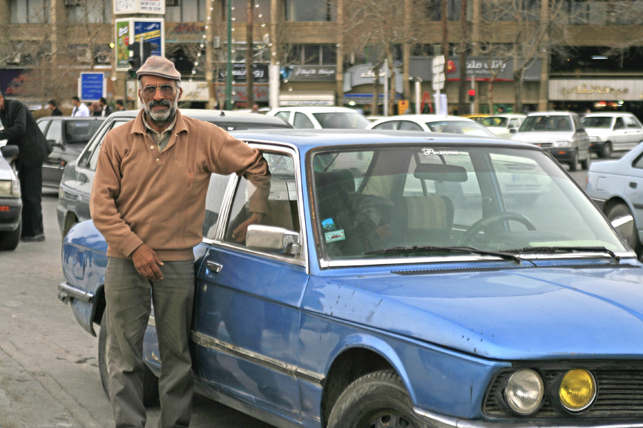 A man in a flat cap leaning on the bonnet of a blue car in a busy street