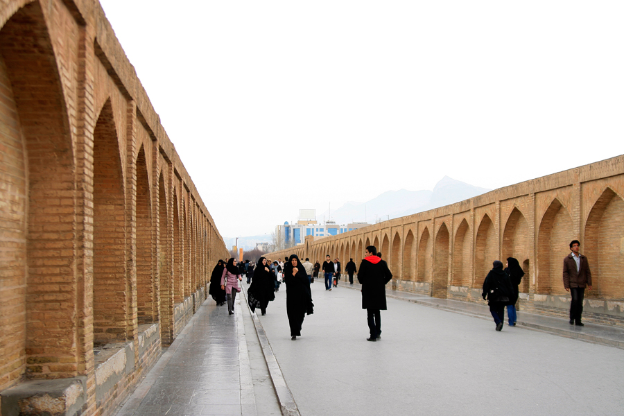 The long brick arcade of a bridge receding into haze, people walking its length