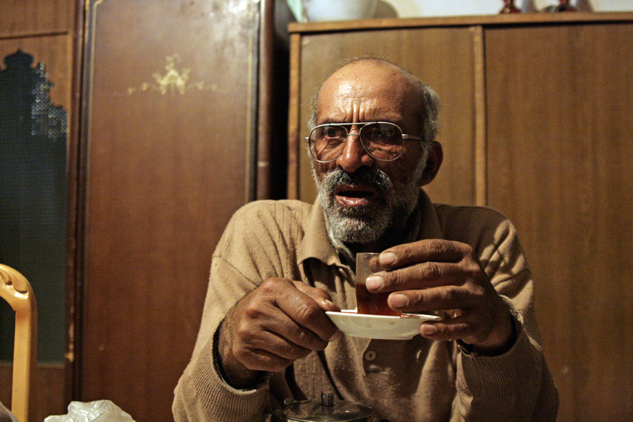 An elderly man in a cardigan drinking tea from a saucer