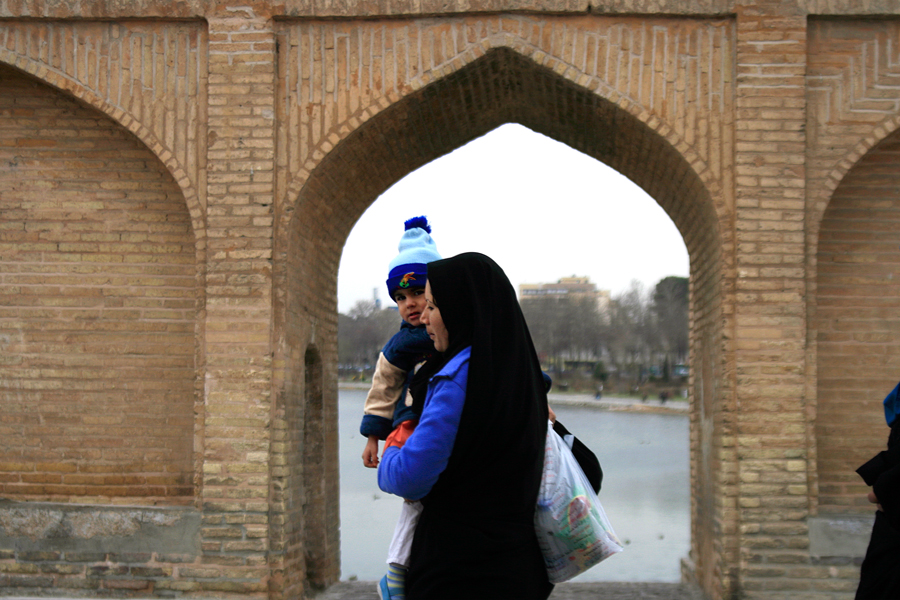 A woman holding a small child in a blue hat, framed in an arch of the bridge, water beyond