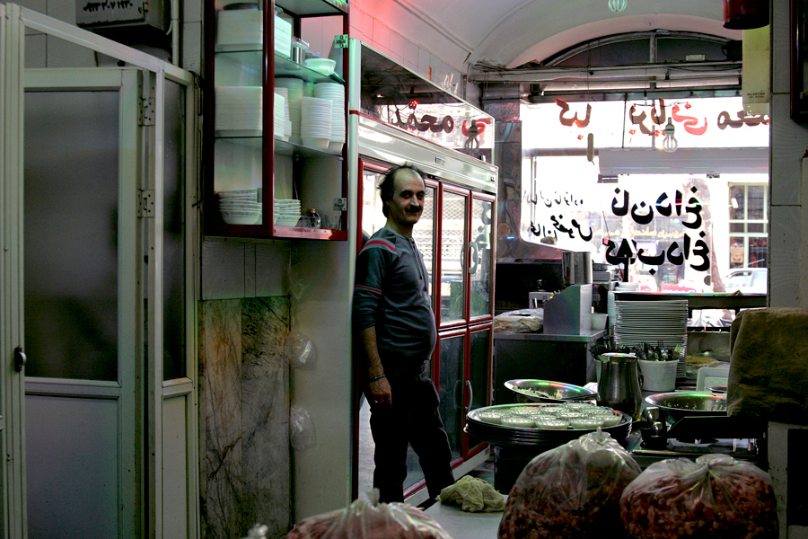 A man standing in his brightly lit food shop, Persian signage above the counter
