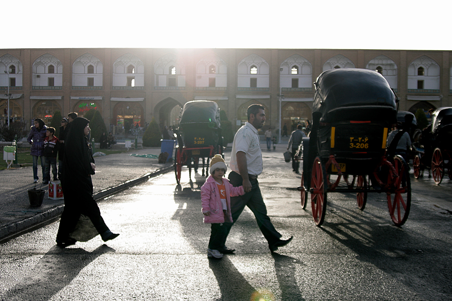 Low sun across a wet square, a family crossing it, horse-drawn carriages waiting
